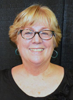 Pam Gannon, smiling, blond, middle aged white woman, wearing a black scoop neck shirt, lasses and earrings