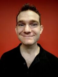 photo of Caleb Lewis, male presenting, short brown hair, wearing a black shirt, standing in front of a red wall